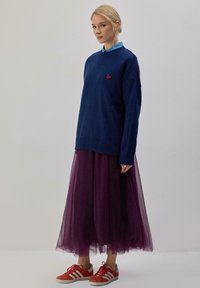 Maglione blu lavorato a maglia con ricamo a cuore rosso, indossato sopra a una lunga gonna di tulle viola trasparente e scarpe sportive con righe rosse.