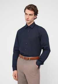 Άνδρας που φοράει μπλε πουκάμισο με κουμπιά, μπαίνει μέσα σε μπεζ παντελόνι με καφέ ζώνη, στέκεται με το ένα χέρι στην τσέπη, κοιτάζει προς τα αριστερά.