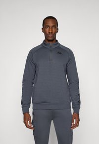 Sudadera gris con cremallera en el cuello, con dobladillo y puños acanalados, detalles del logo en el pecho y las mangas, y una tela suave y texturizada.