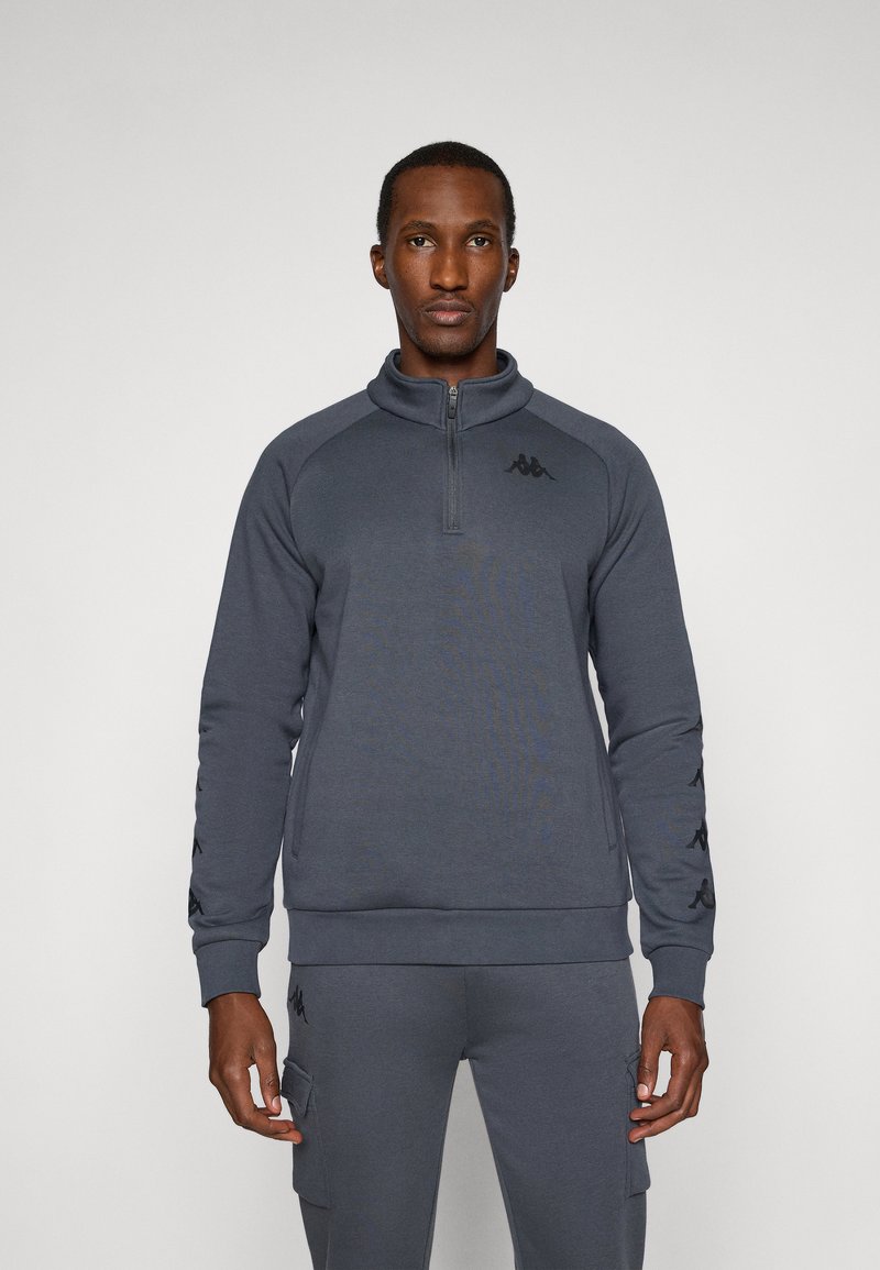 Sudadera gris con cremallera en el cuello, con dobladillo y puños acanalados, detalles del logo en el pecho y las mangas, y una tela suave y texturizada.