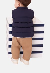 Temno modra puhasta brezrokavnik čez belo majico z dolgimi rokavi, bež hlače in bele superge. Otrok z rinico obrnjeno proti črtasti škatli v belem in temno modrem.