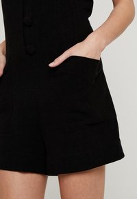 Black linen romper featuring a fitted design, two front pockets, and decorative buttons. Smooth texture and tailored silhouette.