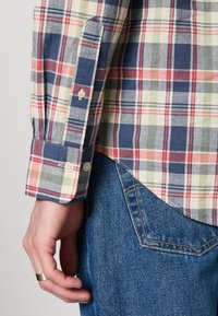 Mannenhand met zilveren ring naast geruite overhemdsmouw met knopen en een achterzak van blauwe spijkerbroek met oranje stiksel.