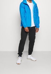 Giacca leggera blu con zip frontale e polsini elastici. Abbinata a pantaloni felpati neri e scarpe sportive bianche. Design semplice.
