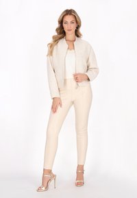 Beige strukturierte Bomberjacke mit Reißverschluss vorne, kombiniert mit cremefarbigen, eng anliegenden Hosen und metallischen Absatzsandalen. Schlichtes Design mit glattem Stoff.