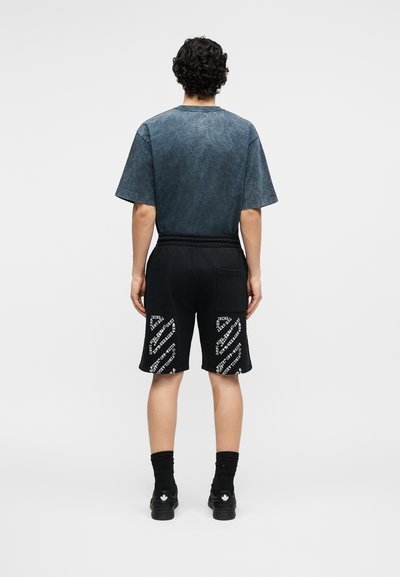 Homme aux cheveux bouclés portant un t-shirt bleu foncé délavé et un short noir avec des motifs diagonaux blancs, debout de dos.