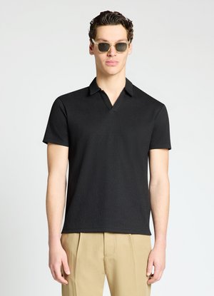 Homme portant des lunettes de soleil monture claire, une chemise noire à manches courtes avec col et un pantalon beige sur fond blanc uni.