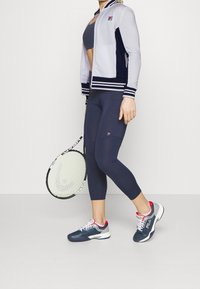 Marina träningsleggings i marinblått, tillsammans med en grå zip-up jacka med marina ränder och en logotyp; kompletterad med grå och röda tennisskor samt ett racket.
