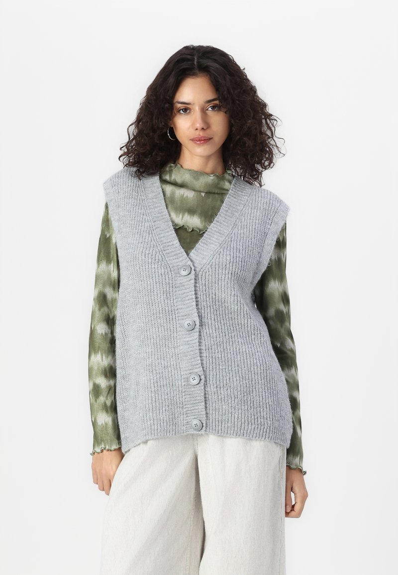 Lichtgrijze gebreide vest met een V-hals, ribbelstructuur en vier knopen. Gelaagd over een groen tie-dye lange mouwtop.