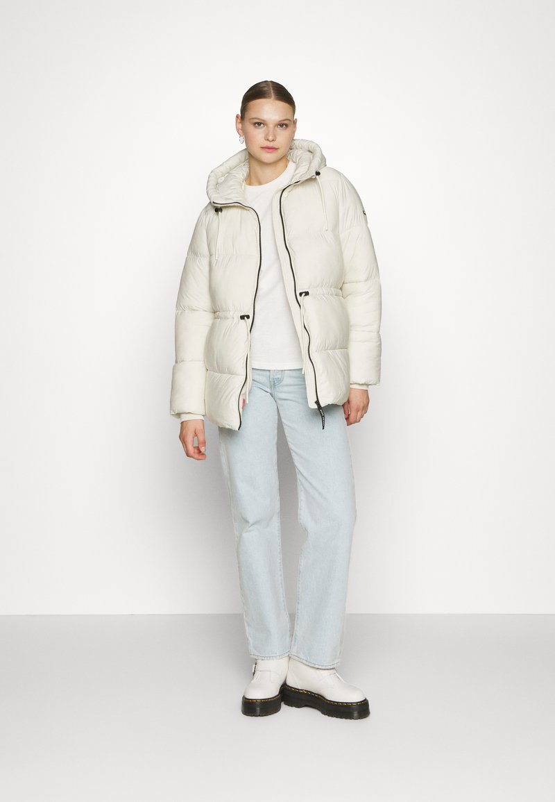 Chaqueta blanca acolchada con capucha, que presenta un cierre de cremallera frontal y detalles de cordón. Lleva puesta sobre una blusa blanca y jeans de color azul claro. Textura suave.