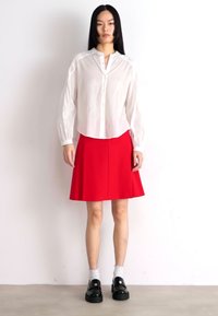 White long-sleeve button-up shirt and red A-line skirt. Black shoes with chunky soles. Plain background, model stands upright.