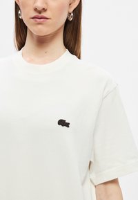 Λευκό βαμβακερό μπλουζάκι με στρογγυλή λαιμόκοψη, με μικρό μαύρο κεντητό λογότυπο στο στήθος. Κοντά μανίκια, άνετη εφαρμογή.