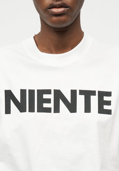 Κοντινό πλάνο ενός ατόμου που φοράει άσπρο πουκάμισο με μαύρο κείμενο "NIENTE" στο στήθος, εμφανή τα χείλη και ο λαιμός, χωρίς άλλα στοιχεία.