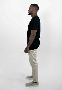 Zwarte t-shirt met korte mouwen gecombineerd met crèmekleurige chinos en grijze instap schoenen. Model staat naar rechts, waardoor de zijaanzicht van de outfit te zien is.