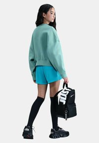 Giovane donna che indossa un ampio felpa teal, pantaloni corti blu, calze nere fino al ginocchio e sneaker, mentre tiene uno zaino Nike nero.