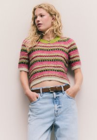 A rövid ujjú, felülvágott pulóver színes csíkos mintákkal díszített rózsaszín, zöld és fekete színekben, texturált kötött anyagból készült. Világoskék farmerrel viselve.