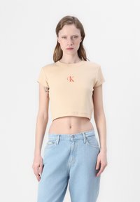 Korte beige t-shirt van ribstof met korte mouwen, voorzien van een contrasterend oranje 'CK' logo op de voorkant. Gecombineerd met lichtblauwe jeans.