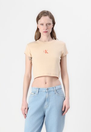 Cropped beige t-shirt made of ribbed fabric with short sleeves, featuring a contrasting orange 'CK' logo on the front. Paired with light blue jeans.