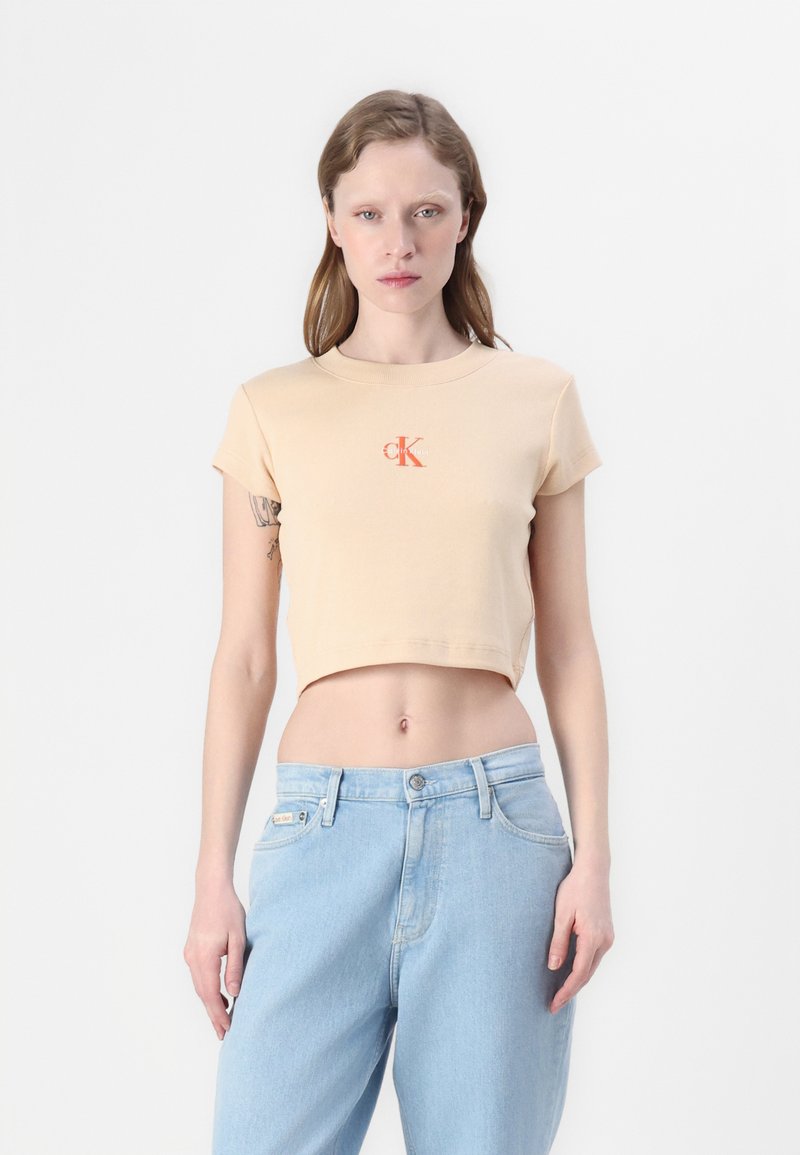 Korte beige t-shirt van ribstof met korte mouwen, voorzien van een contrasterend oranje 'CK' logo op de voorkant. Gecombineerd met lichtblauwe jeans.