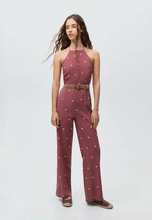 Red polka dot jumpsuit with a halter neck design, belted at the waist with a woven belt, paired with brown flat shoes. Textured fabric.