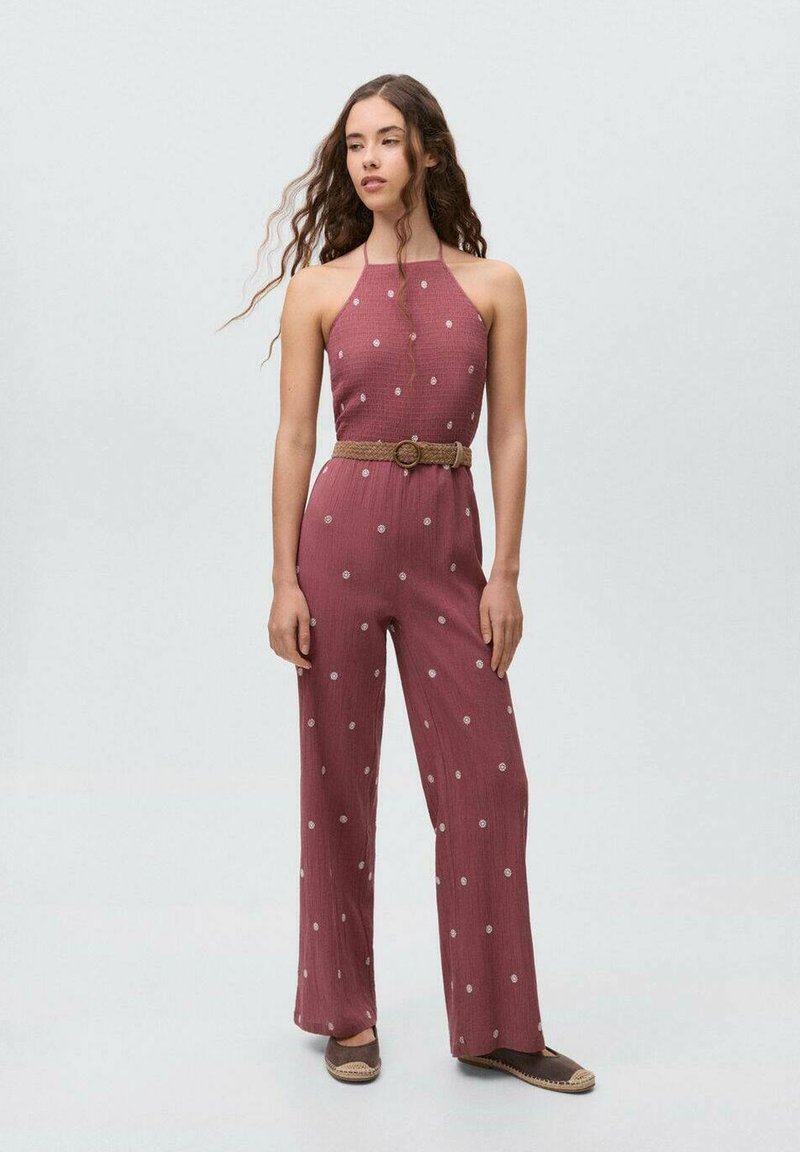 Red polka dot jumpsuit with a halter neck design, belted at the waist with a woven belt, paired with brown flat shoes. Textured fabric.