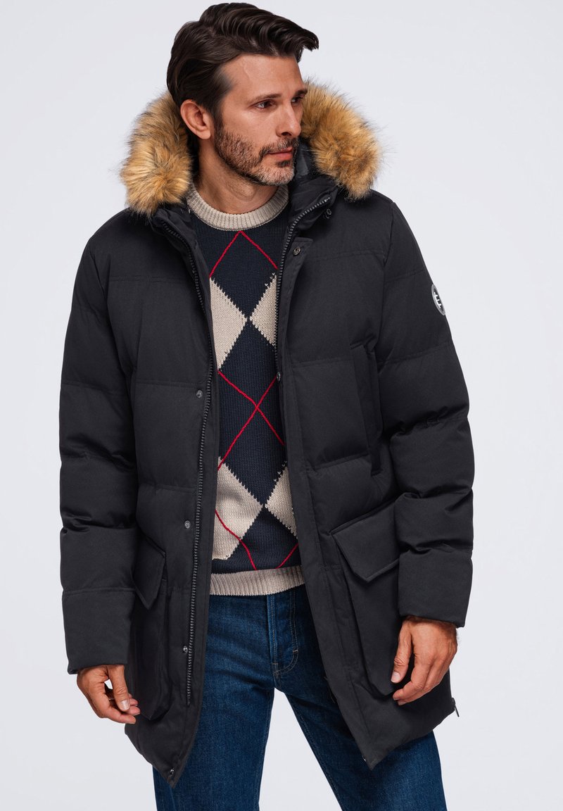 Homme portant un parka d’hiver noir avec capuche en fourrure, un pull à losanges et un jean bleu, regardant vers sa gauche sur un fond uni.
