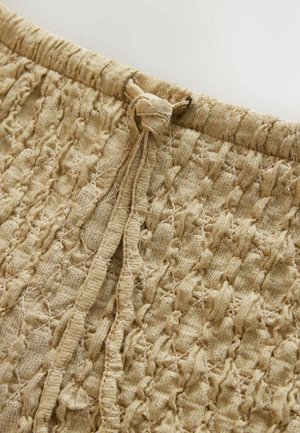 Teksturisana bež tkanina s naboranim dizajnom, koja ima skupljeni pojas i vezani detalj. Materijal izgleda lagano i prozračno.