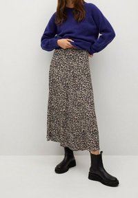 Donna che indossa un maglione viola, una lunga gonna nera con fiori e stivaletti neri, in piedi contro uno sfondo chiaro e uniforme.