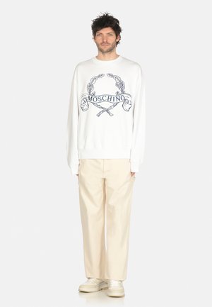 Felpa bianca con logo Moschino e design grafico, abbinata a pantaloni beige chiaro a gamba larga e sneakers beige.