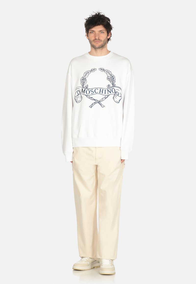 Felpa bianca con logo Moschino e design grafico, abbinata a pantaloni beige chiaro a gamba larga e sneakers beige.