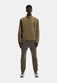 Helle braune Halb-Reißverschlussjacke mit elastischen Bündchen, kombiniert mit grauen schmalen Hosen und weißen Sportschuhen. Das Outfit hat ein minimalistisches Design.