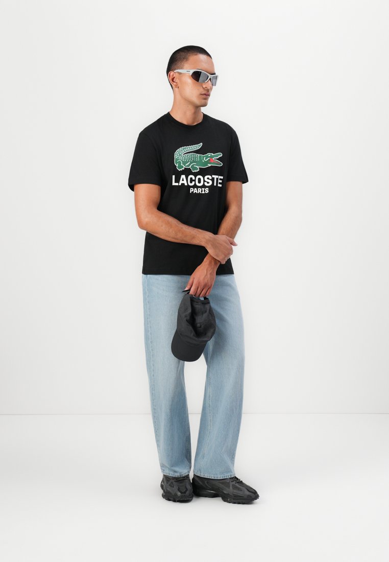 Черен памучен потник с зелено крокодилско лого и бял текст "LACOSTE PARIS"; светлосини дънки; черна шапка и маратонки; сребърни слънчеви очила.