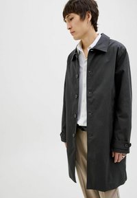 Palton lung, negru, cu un design clasic, având butoane mari, guler ascuțit și detalii la manșete. Purtați peste o cămașă albă și pantaloni bej.