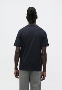 Mees lühikeste dredsidega, musta t-särgi ja hallide teksadega, seisab seljaga valgele lihtsale taustale.