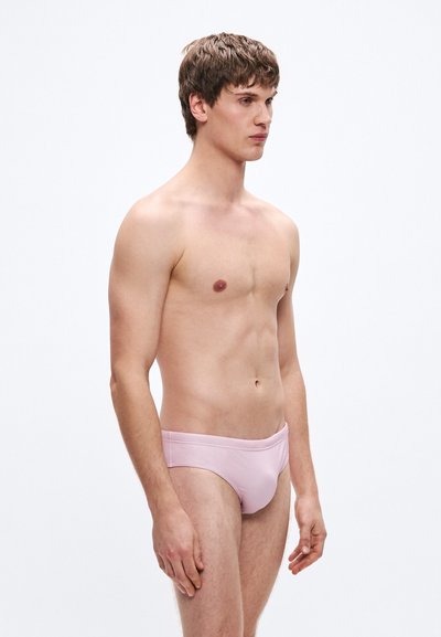 Heledad roosa ujumispüksid, millel on sile ja elastne kangas ning keskmise kõrgusega lõige, mida kannab paljaste ülakerega meesmodell profiilis.