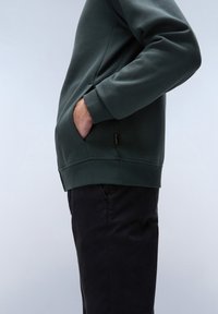 Sudadera verde oscura con puños y dobladillo de canalé, que cuenta con bolsillos laterales y un corte relajado. Combinada con pantalones oscuros para un look casual.