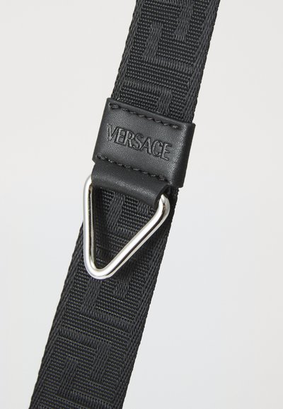Black fabric strap with embossed geometric pattern, leather detail stamped with "VERSACE," and a silver triangular metal ring.