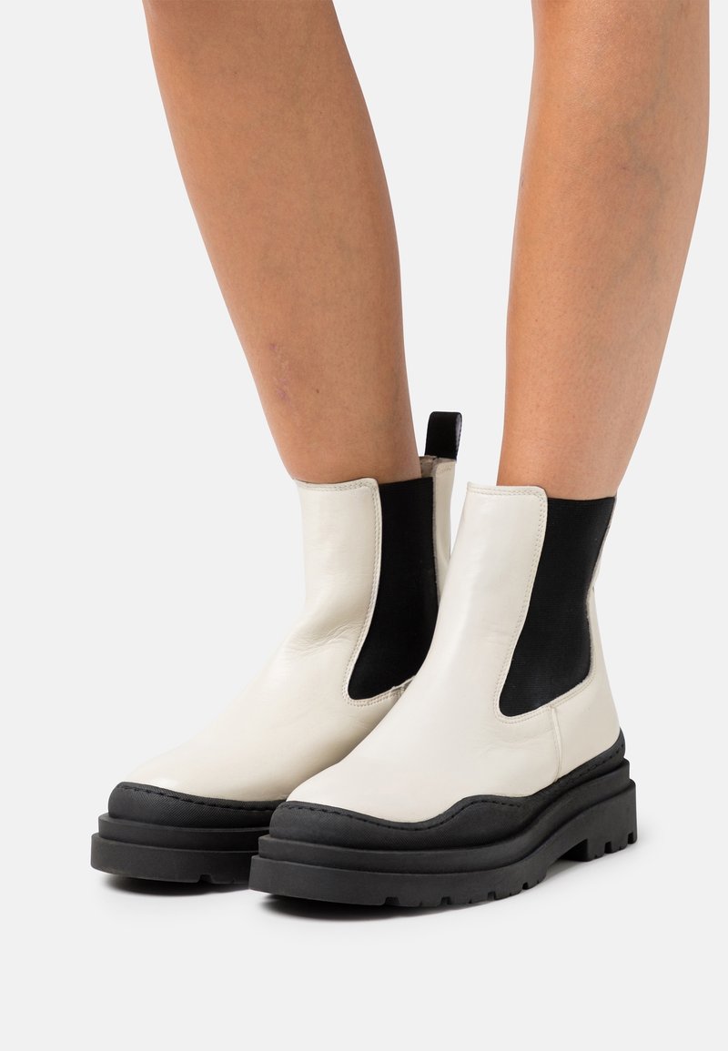 Bottes blanches en cuir à la cheville avec panneaux latéraux élastiques noirs et semelle épaisse en caoutchouc noir. Bout arrondi avec détails de couture contrastants.
