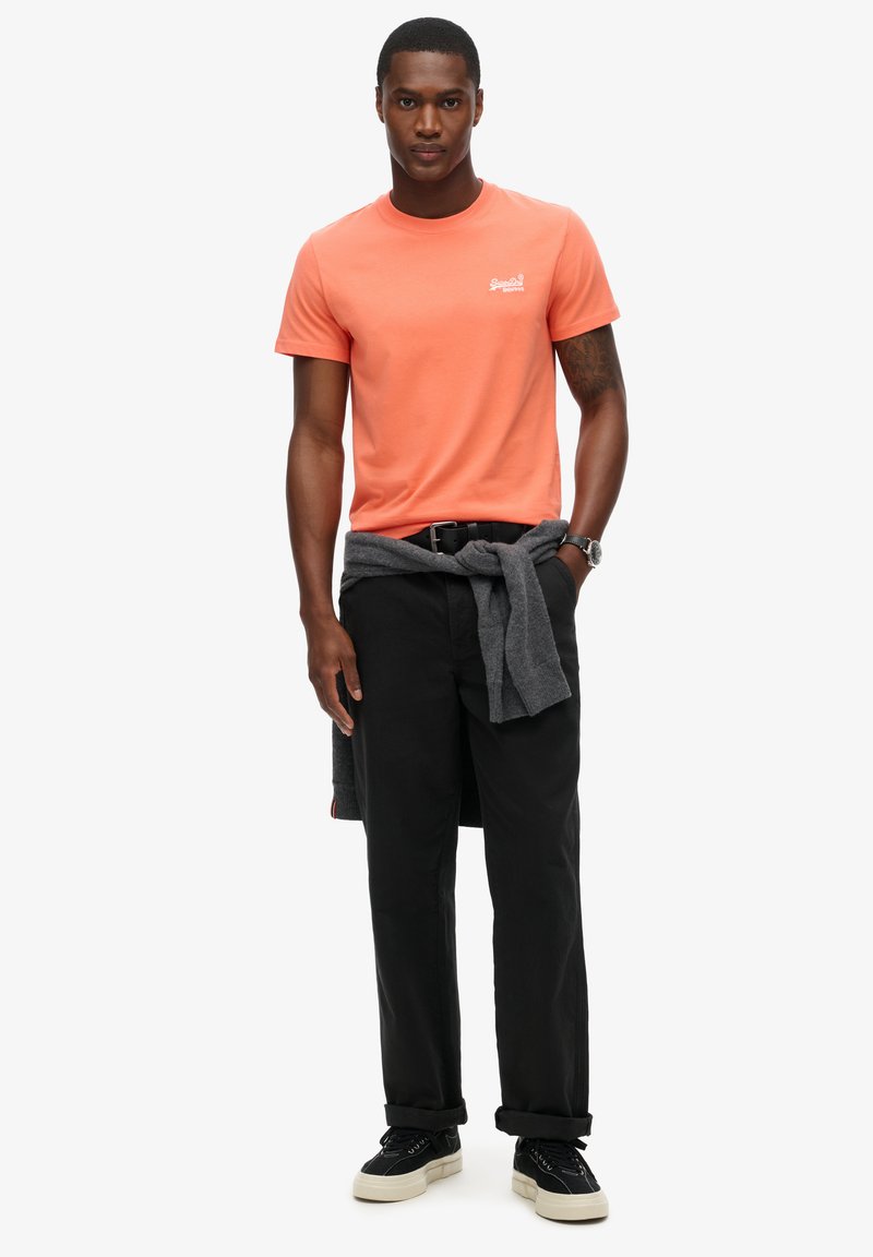 T-shirt in cotone corallo con scollo rotondo e logo, abbinato a pantaloni neri e un maglione grigio scuro annodato in vita. Sneakers nere ai piedi.