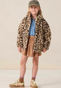 Jong meisje in een panterprint jas en bijpassende pet, spijkerblouse, tan rok, bruine gebreide sokken en beige sneakers staat zelfverzekerd.