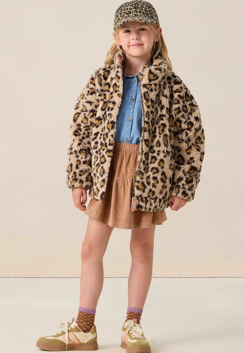 Jong meisje in een panterprint jas en bijpassende pet, spijkerblouse, tan rok, bruine gebreide sokken en beige sneakers staat zelfverzekerd.