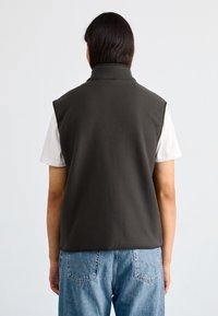 Dunkelgraue Fleece-Weste mit hohem Kragen, ärmellosem Design und glatter Textur, kombiniert mit blauen Jeans. Schlichter und funktionaler Stil.
