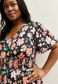 Mujer vestida con un vestido negro con patrón floral en rosa y verde, cintura elástica, mangas cortas y un delicado collar de oro.