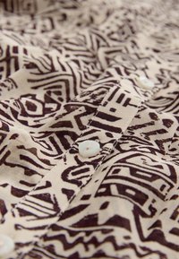 Button-up shirt with a beige fabric featuring a dark brown abstract pattern. Notable details include white buttons and stitched seams.