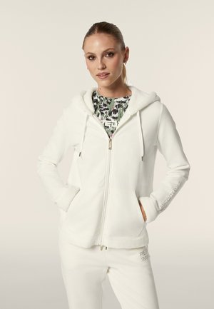 Haut à zip blanc avec une capuche douce et douillette, présentant une texture lisse, des poches avant et un logo brodé sur la manche.