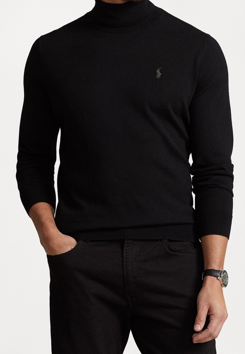Homme portant un pull à col roulé noir à manches longues avec un petit logo brodé sur la poitrine et un pantalon noir.