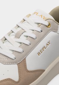 Λευκό και μπεζ αθλητικό παπούτσι με λευκά κορδόνια, λεπτομέρεια χρυσής τρύπας για το κορδόνι και το λογότυπο "REPLAY" στην πλευρά και τη γλώσσα.