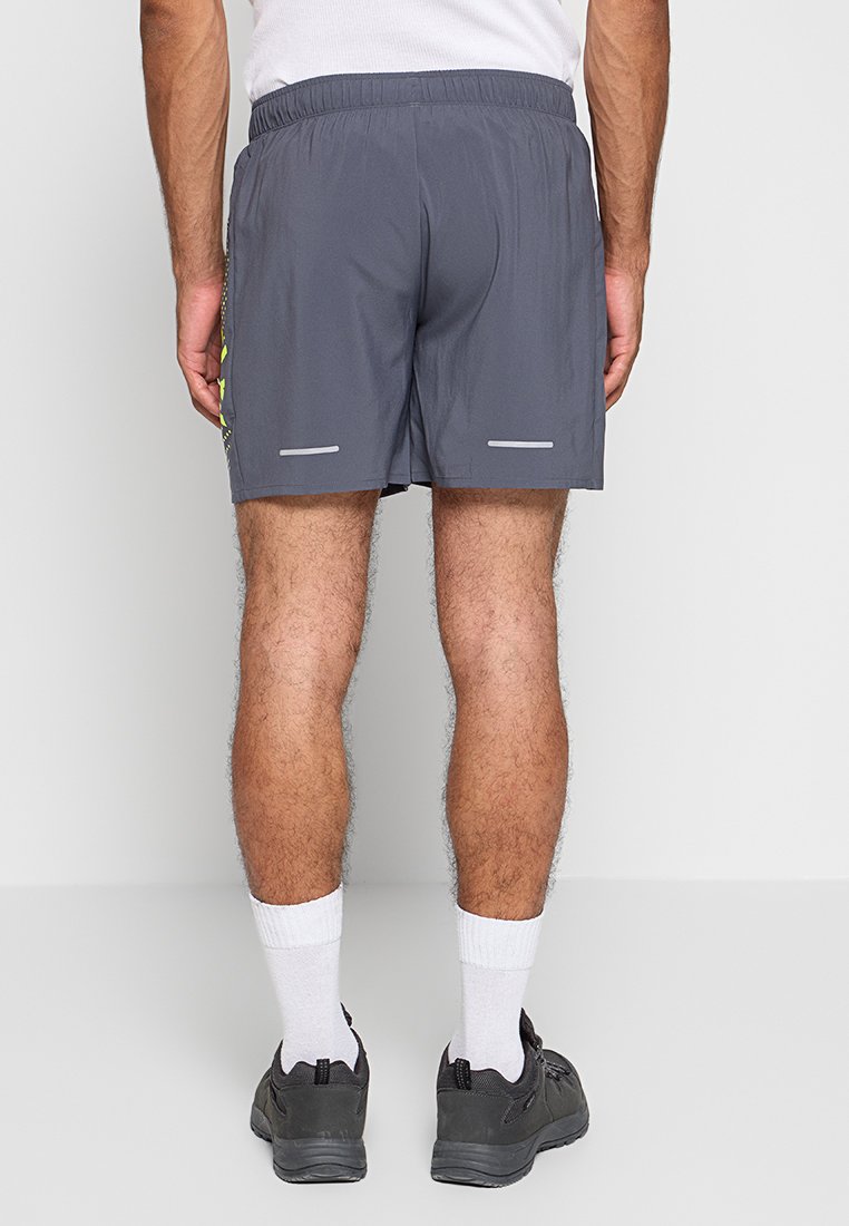 Man in grijze sportshorts, witte sokken en zwarte sneakers, met de rug naar de camera gekeerd tegen een effen achtergrond.