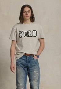 Mladý muž s vlasy po ramena, oblečený v béžovém tričku "POLO", roztrhaných modrých džínách, hnědém opasku a modrých hodinkách, stojící na jednolitém pozadí.