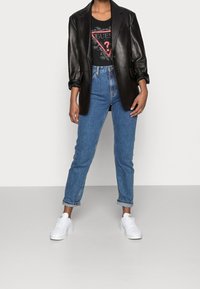 Svart läderblazer över en svart grafisk t-shirt med en triangulär design, ihopparat med ljusblå högmidjade jeans och vita sneakers.